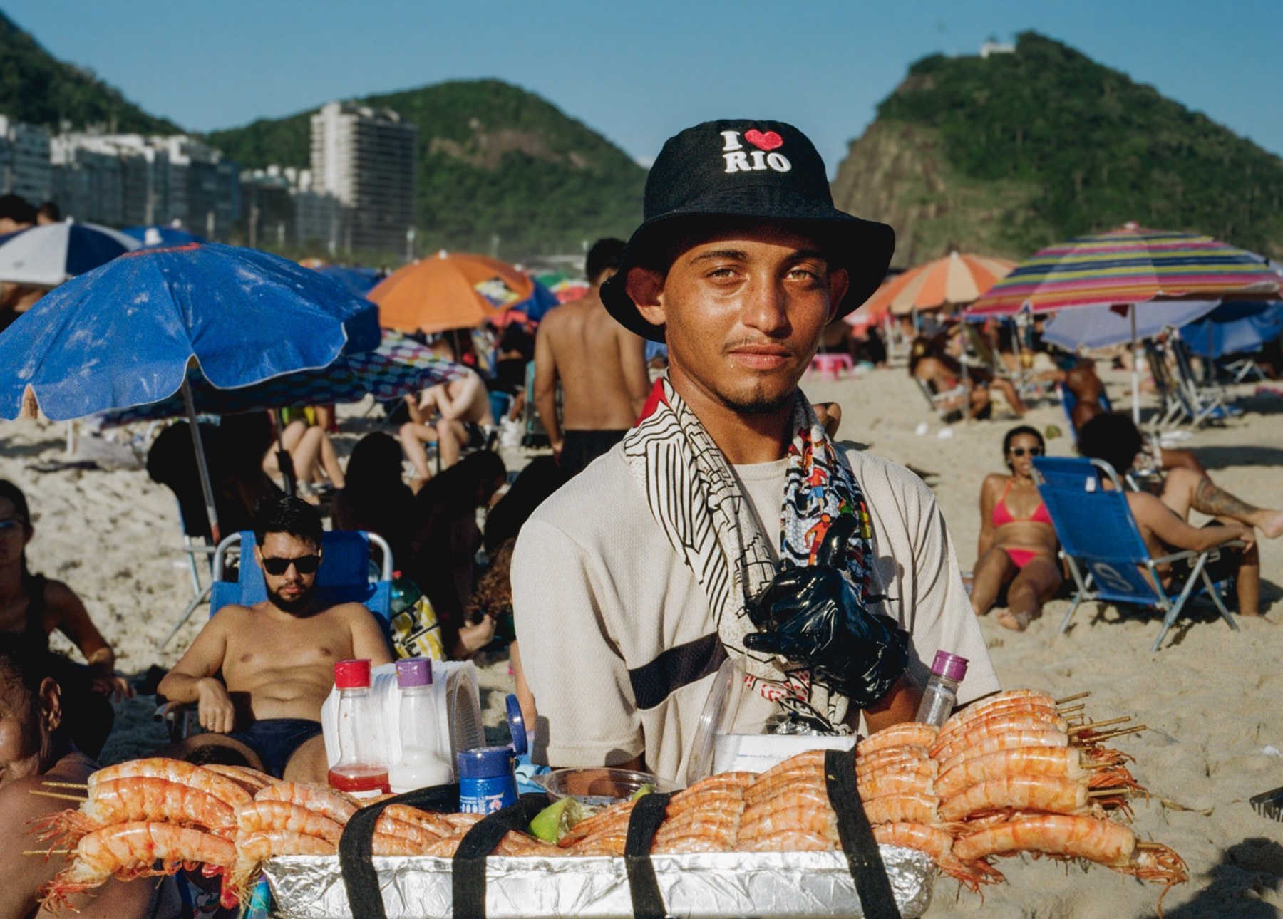 Portrait of a beach vendor in an I love Rio bucket hat holding a tray of grilled prawns