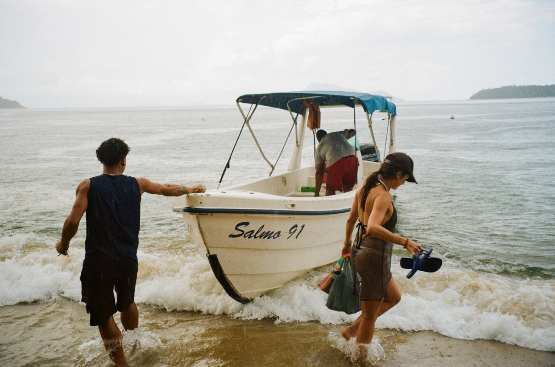 Two people pushing a small boat named Salmo 91 into the surf in the rain