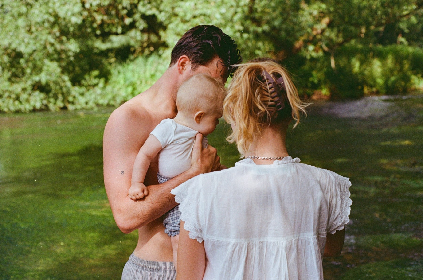 A man holds a baby beside a woman in a white dress by a green river