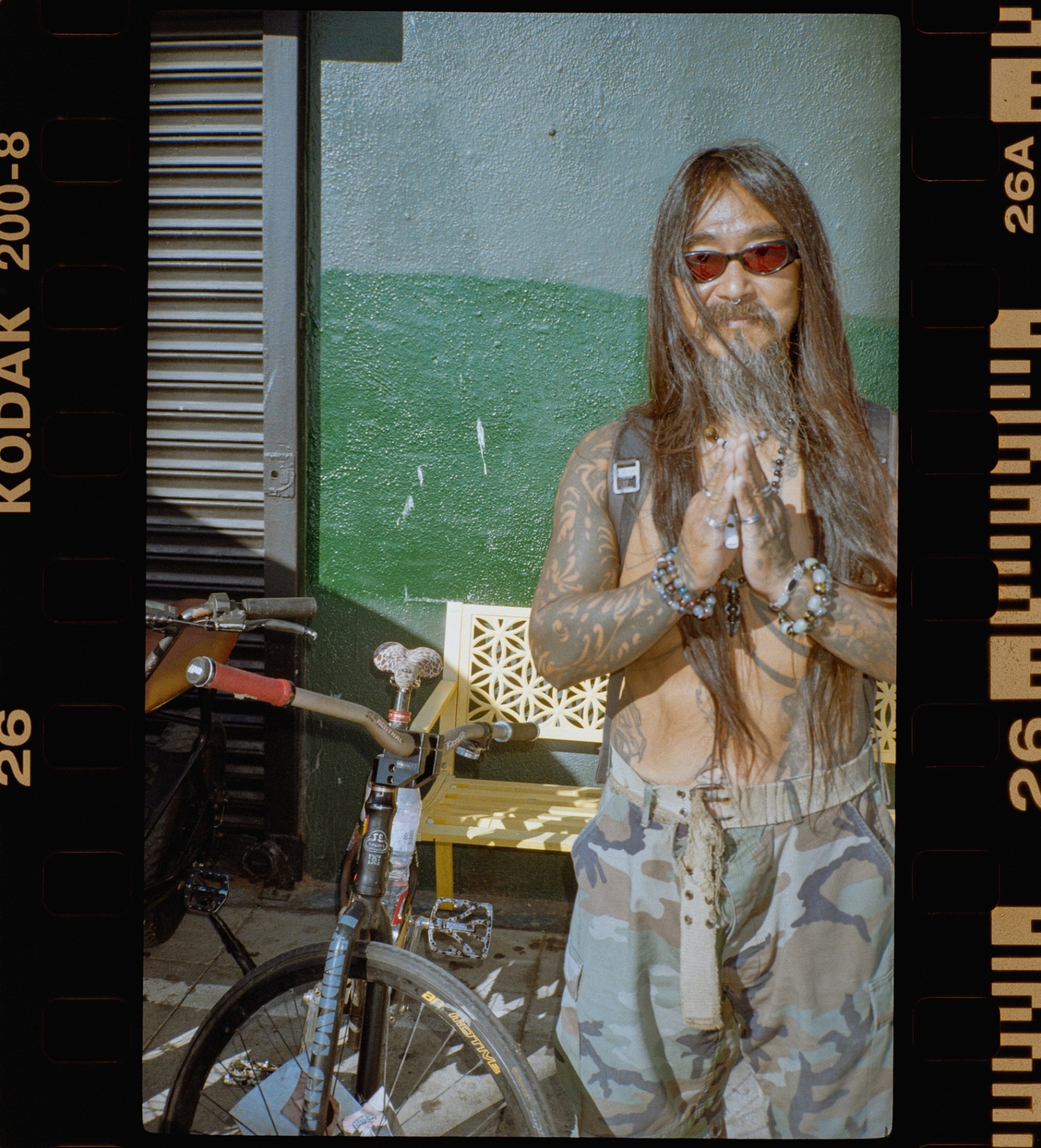 Long-haired man with tattoos and sunglasses standing beside a bicycle, hands pressed together